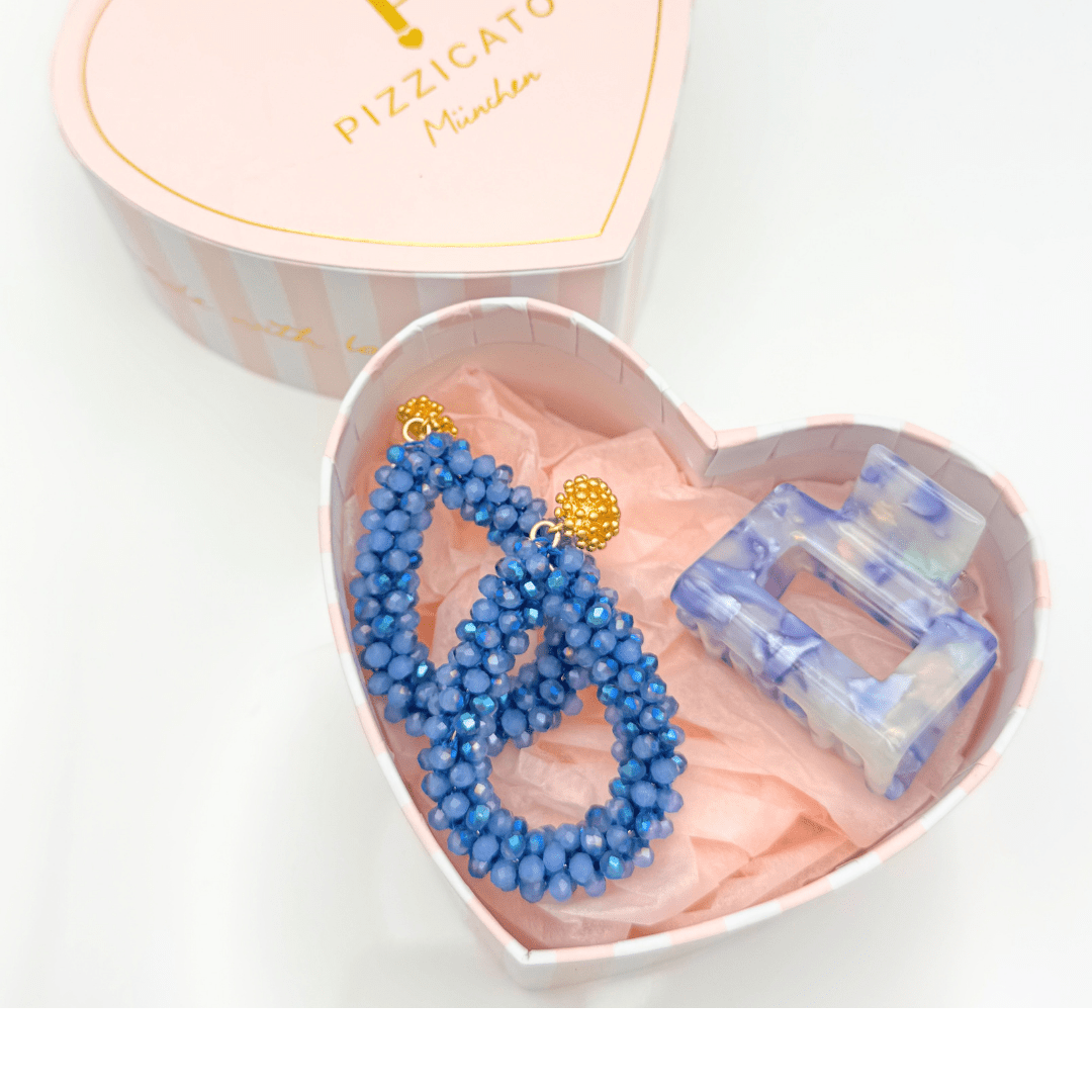 Geöffnete Pizzicato Herzgeschenkbox mit blauen Statement-Ohrringen und hellblauer Haarspange